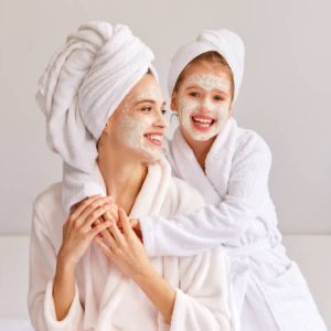 Cheerful girl in bathrobe and towel smiling and embracing young woman with moisturizing mask while resting on bed at home